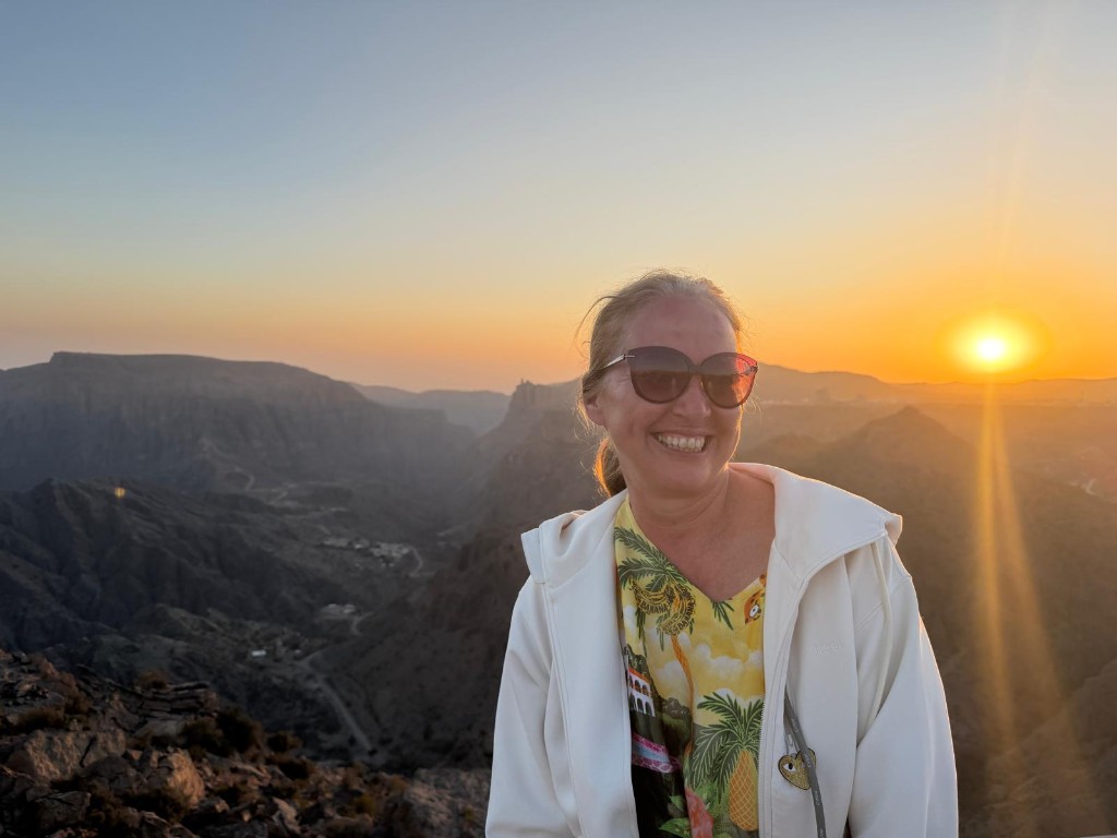 Maria Baumgartner in weißer Jacke und Sonnenbrille, lächelnd vor Berglandschaft im Abendlicht.