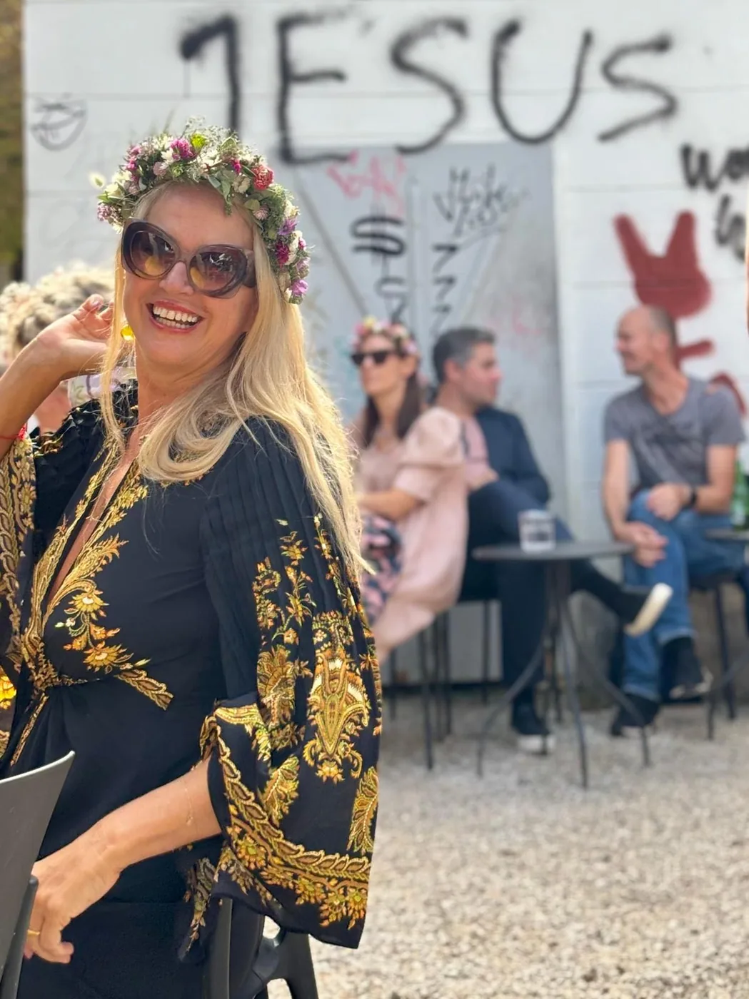 Maria mit Blumenkranz und gemustertem Mantel vor einer urbanen Wand.